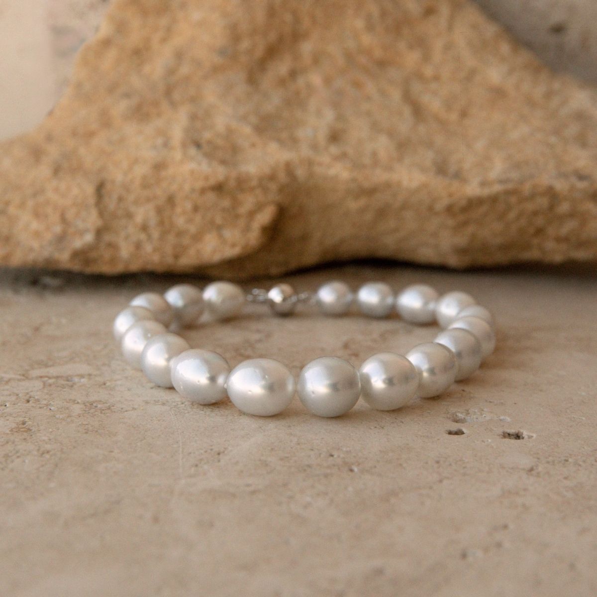 A pearl strand bracelet made of large, lustrous white South Sea pearls with a small 9ct white gold ball clasp, displayed on a textured beige stone surface with a larger rock in the background.