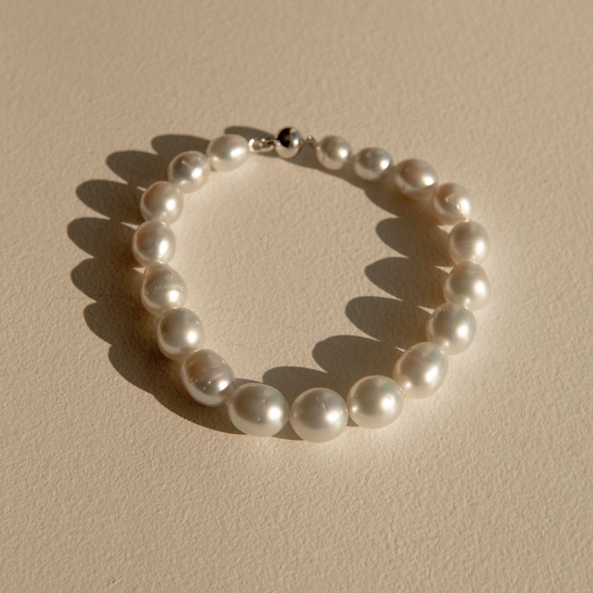 A strand of white silver toned Australian South Sea pearls arranged in a circle on a textured beige surface, with a silver-toned clasp visible at the top and soft shadows cast to the left.