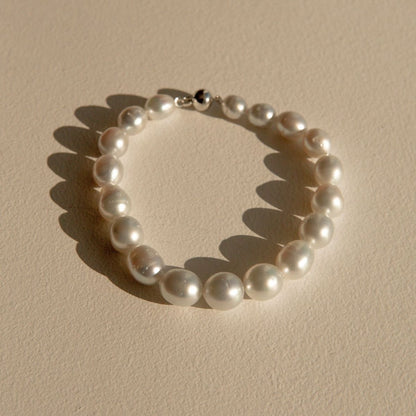 A strand of white silver toned Australian South Sea pearls arranged in a circle on a textured beige surface, with a silver-toned clasp visible at the top and soft shadows cast to the left.