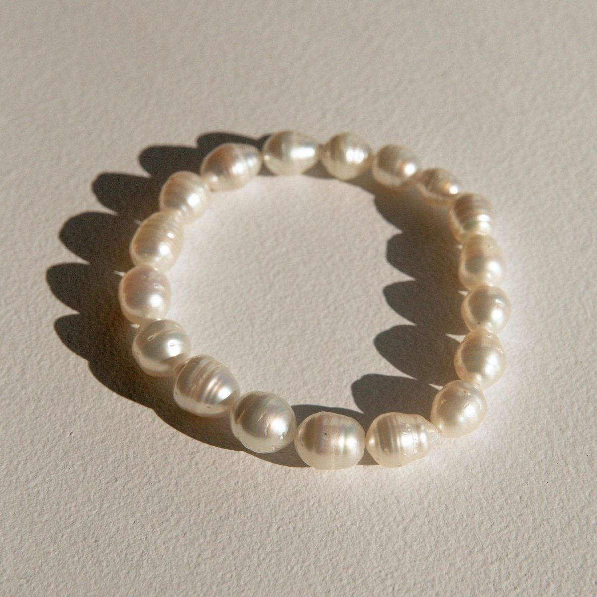 A strand bracelet made of Australian South pearls, arranged in a circle on a textured beige surface with a hard shadow cast to the left.