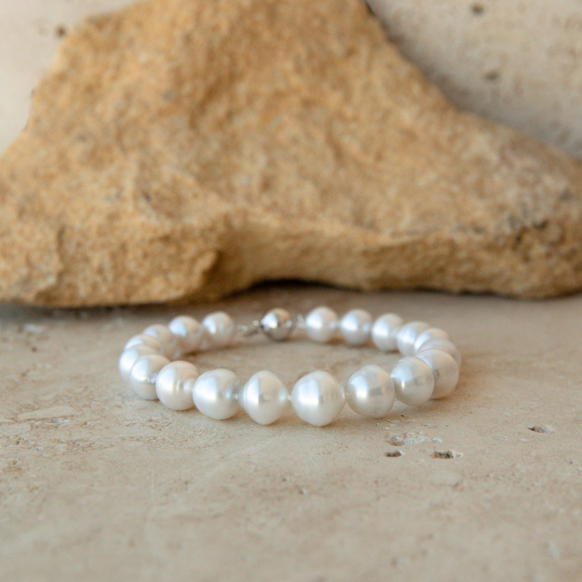 A strand bracelet made of white Australian South Sea pearls, displayed on a textured beige stone surface with a larger rock in the background.