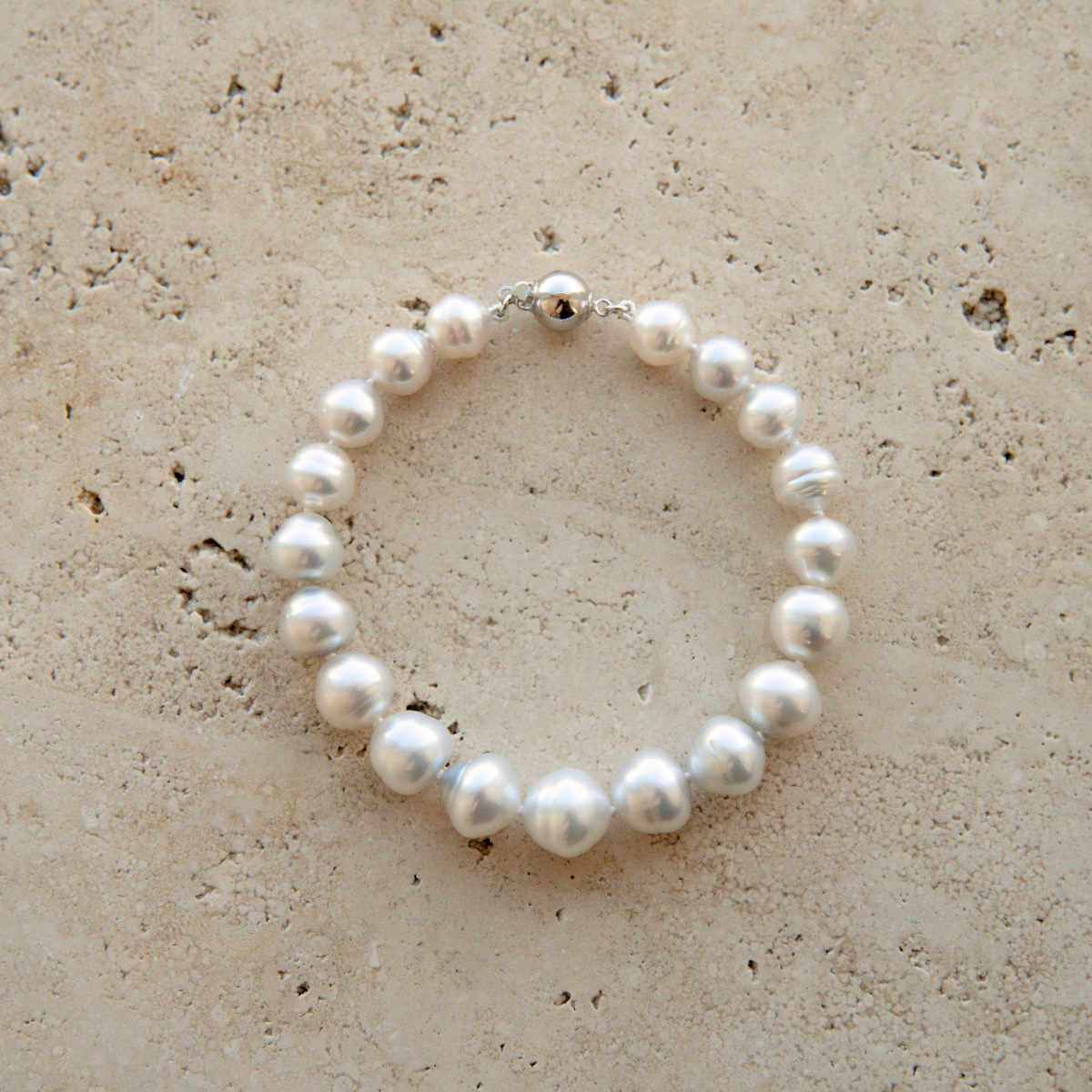 An Australian South Sea pearl bracelet with a silver clasp, arranged in a circle on a textured beige stone surface.