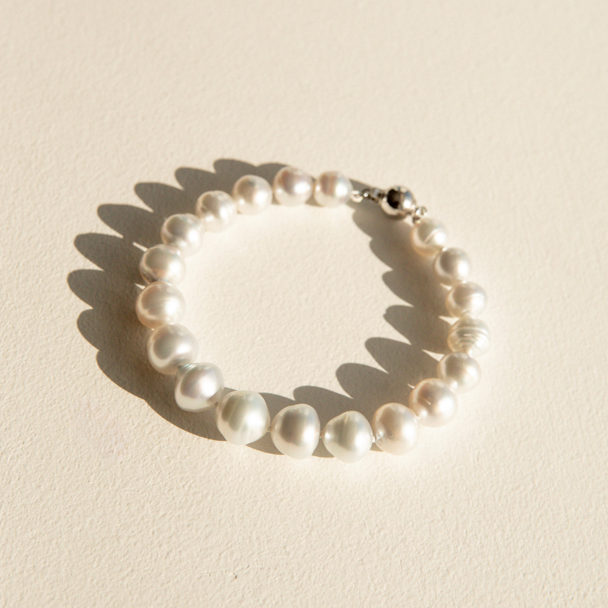 A strand bracelet made of irregular white Australian South Sea pearls with a silver-toned clasp, photographed on a warm off-white textured surface with a soft shadow.