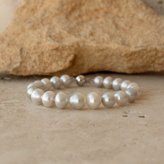 A pearl strand bracelet made of irregular white Australian South sea pearls with a 9ct white gold ball clasp, resting on a textured beige stone surface with a larger tan rock in the background.