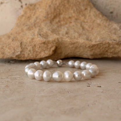 A pearl strand bracelet made of round white South Sea pearls with a 9ct white gold ball clasp, displayed on a textured stone surface in front of a larger rock.