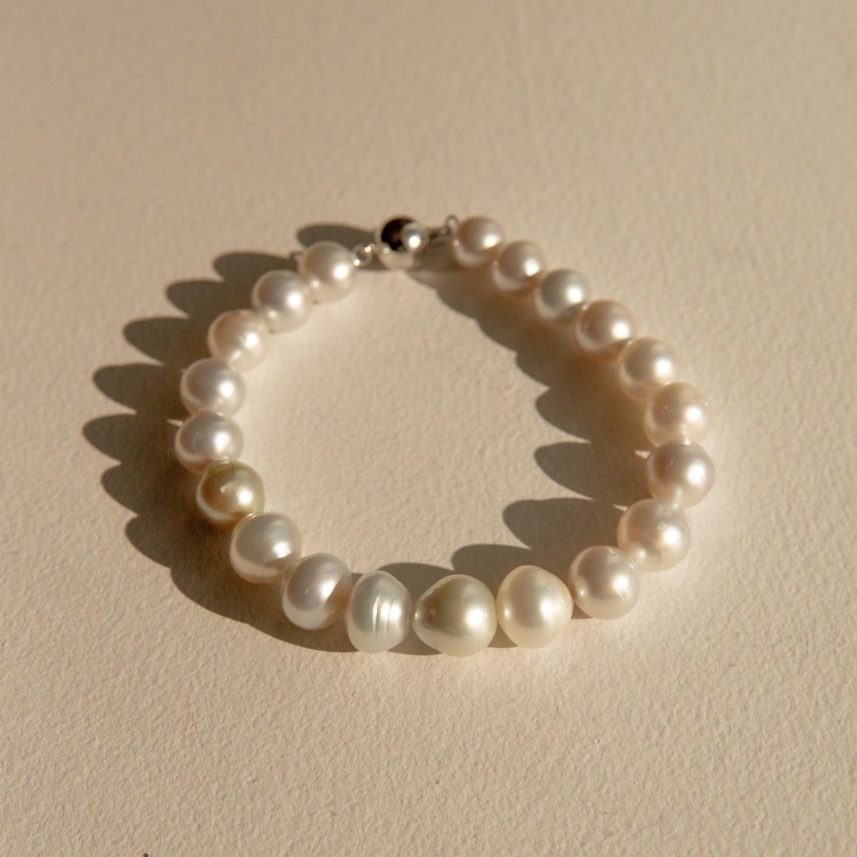 A Saltwater pearl strand bracelet with a 9ct white gold ball clasp, arranged in a circle on a textured beige surface with soft shadows.
