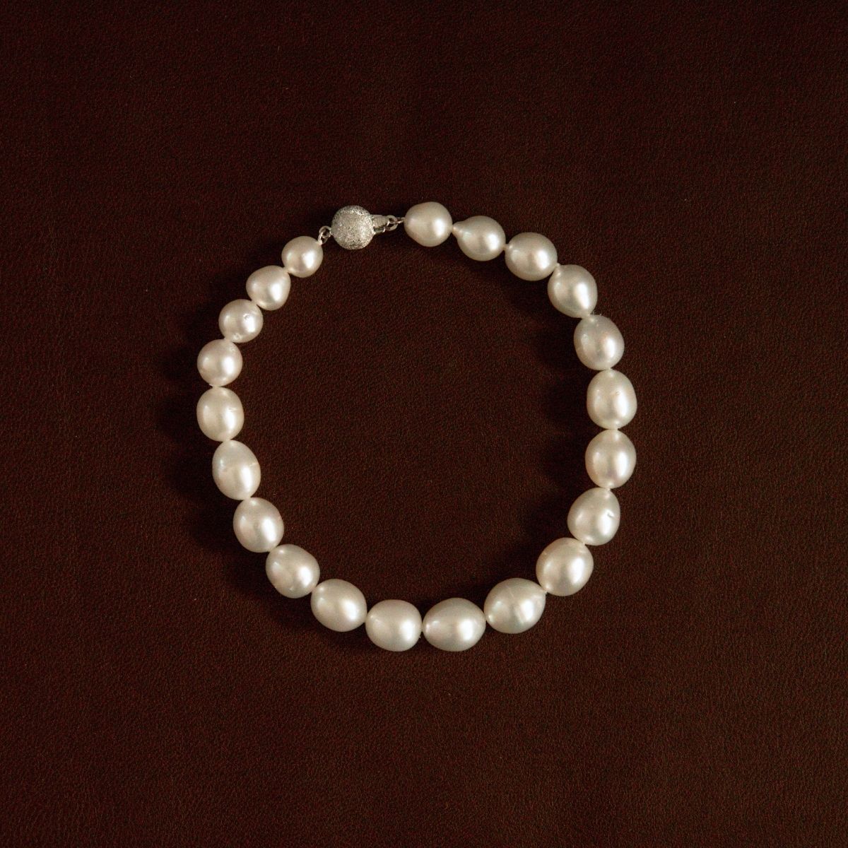 A classic pearl strand necklace arranged in a circle on a dark brown textured background, featuring a silver-toned clasp with a clear crystal accent.
