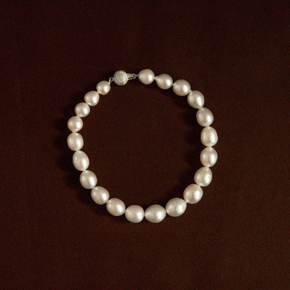 A classic pearl strand necklace arranged in a circle on a dark brown textured background, featuring a silver-toned clasp with a clear crystal accent.