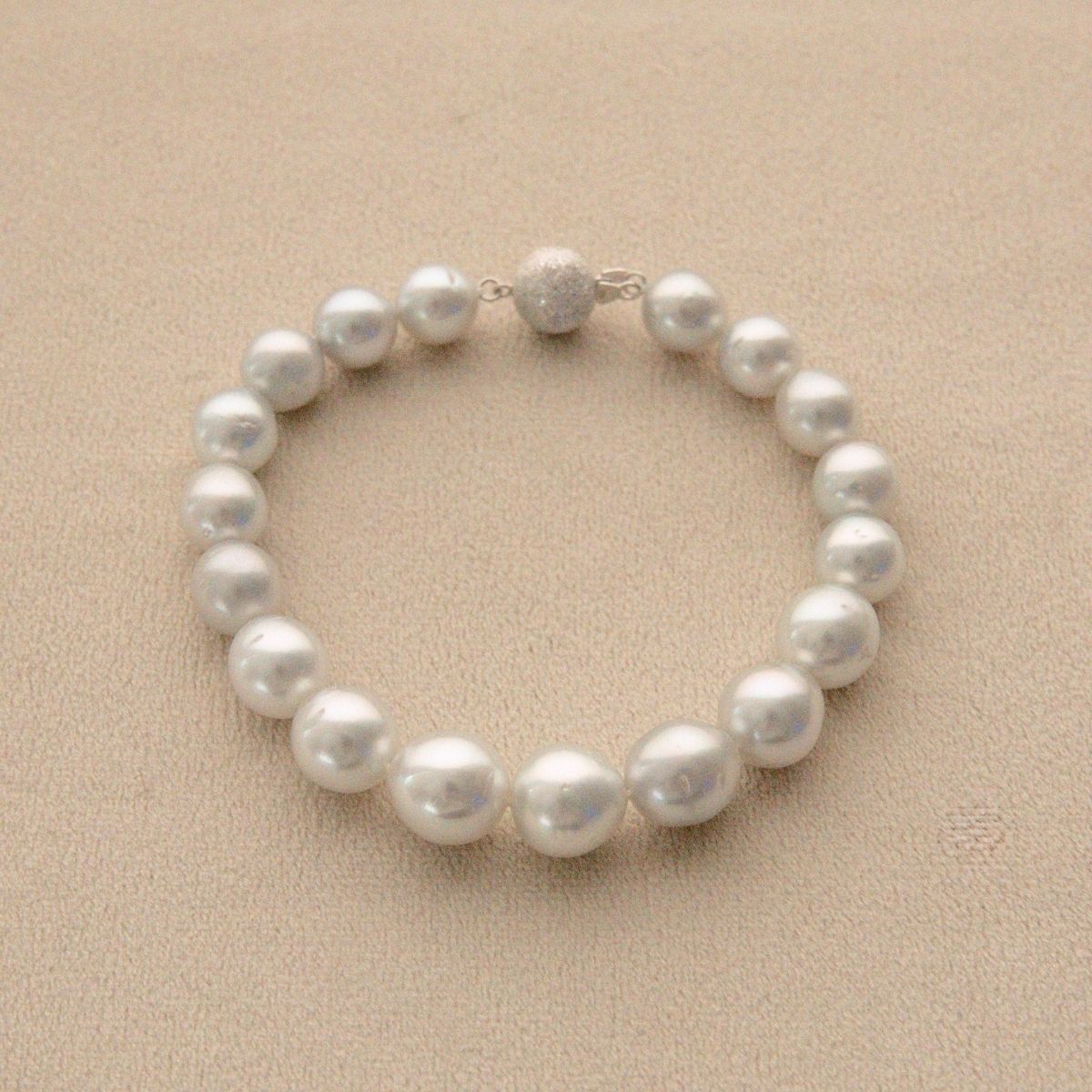 A strand bracelet made of round white Australian South Sea pearls with a textured white gold clasp, displayed on a textured beige background.
