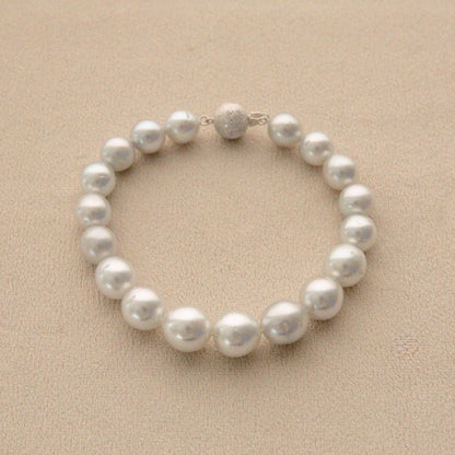 A strand bracelet made of round white Australian South Sea pearls with a textured white gold clasp, displayed on a textured beige background.