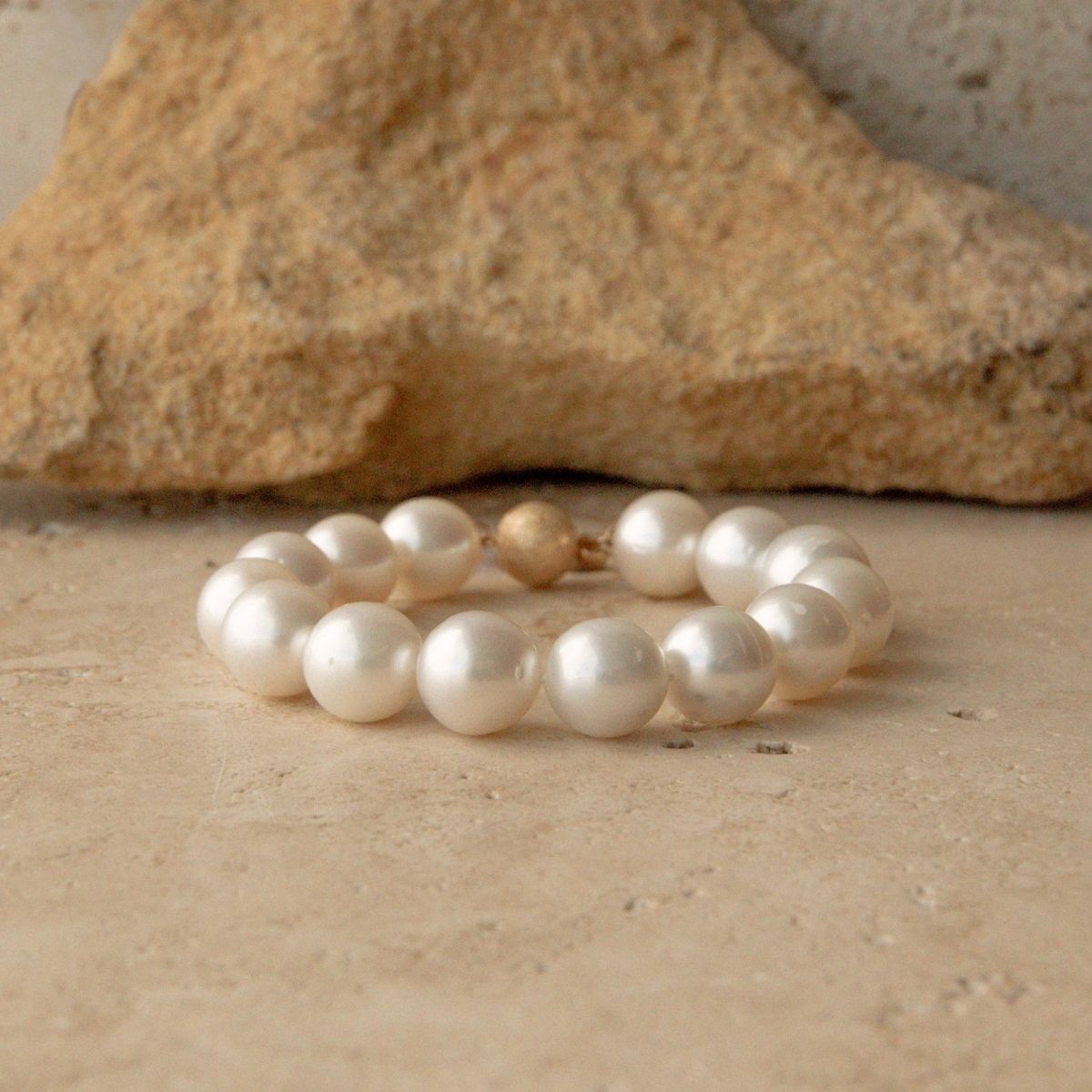 A pearl strand bracelet made of white  Australian South Sea pearls with a single small metallic 9ct yellow gold ball clasp, displayed on a beige stone surface with a larger rock in the background.