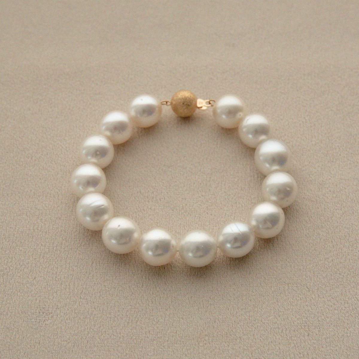 An Australian South Sea pearl strand bracelet with a textured 9ct yellow gold clasp, displayed on a neutral beige textured background.