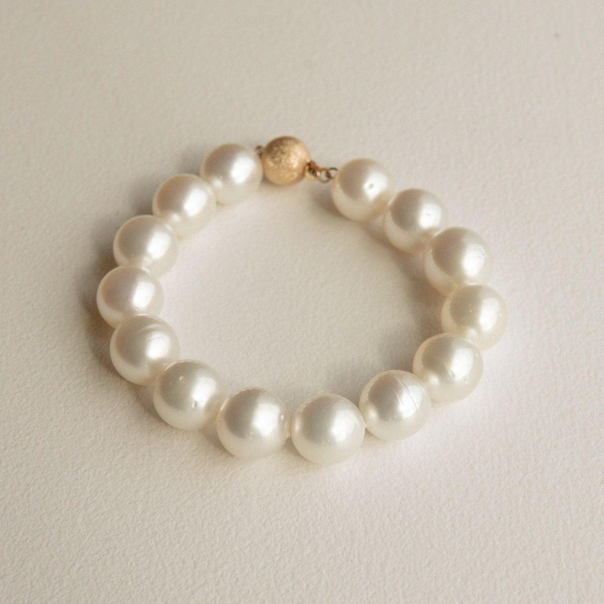 An Australian South Sea pearl strand bracelet made of large, lustrous white Saltwater pearls with a small textured 9ct yellow gold clasp, displayed on a neutral cream-colored surface.