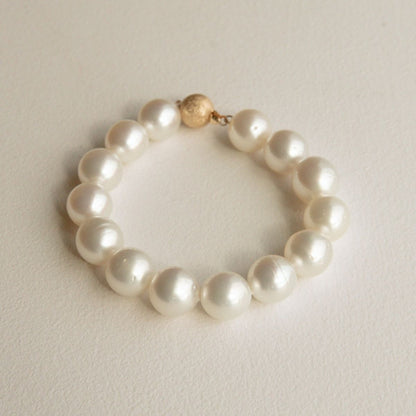 An Australian South Sea pearl strand bracelet made of large, lustrous white Saltwater pearls with a small textured 9ct yellow gold clasp, displayed on a neutral cream-colored surface.