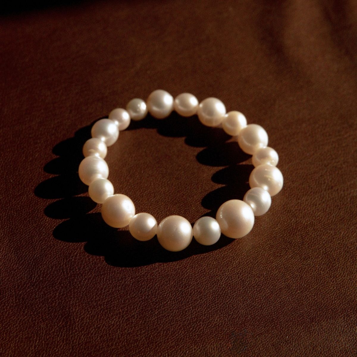 A pearl strand bracelet made of large, lustrous white Cygnet Bay grown Australian South Sea pearls, arranged in a circle on a dark brown textured leather background.