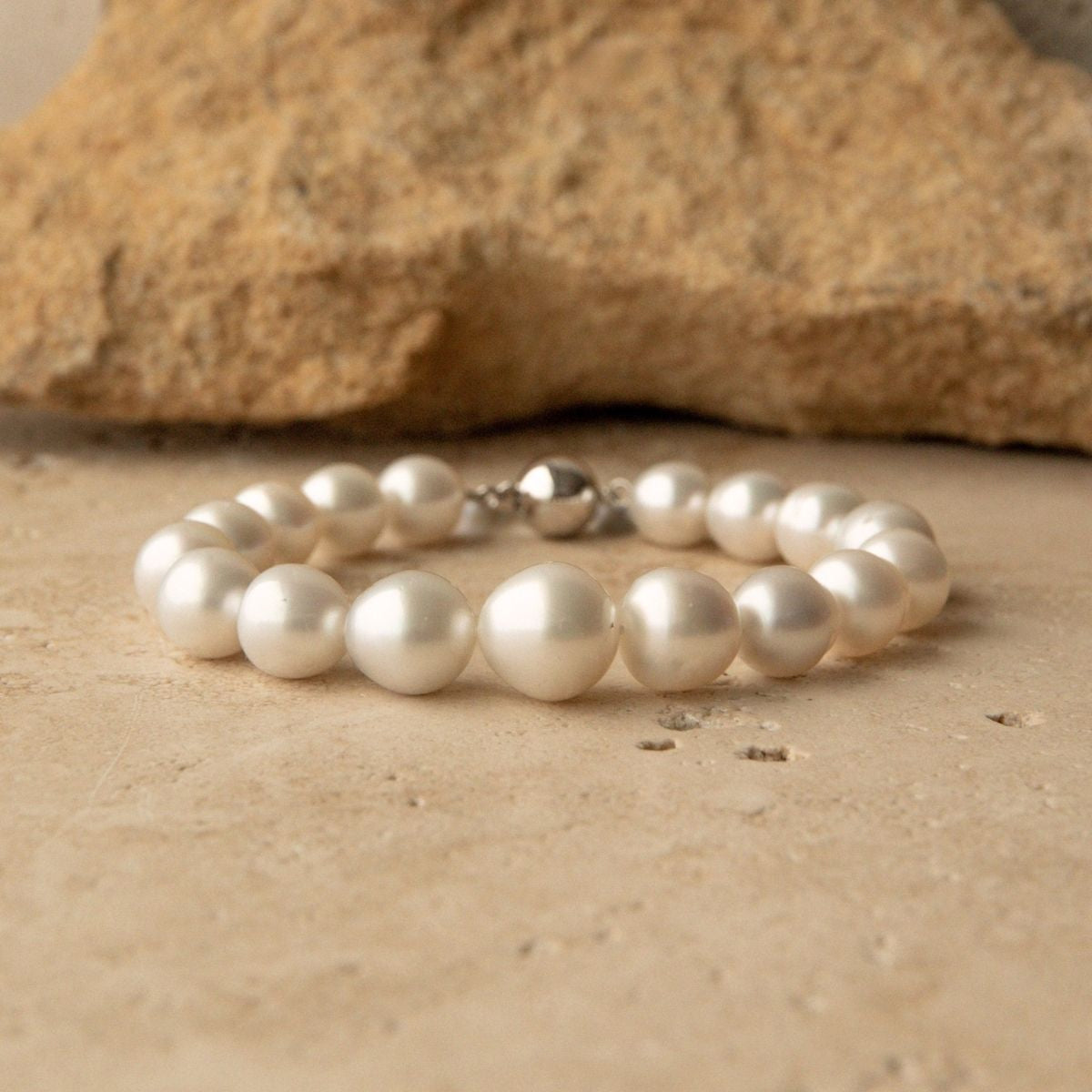 A pearl strand bracelet with a 9ct white gold ball clasp, displayed on a textured beige stone surface with a larger rock in the background.