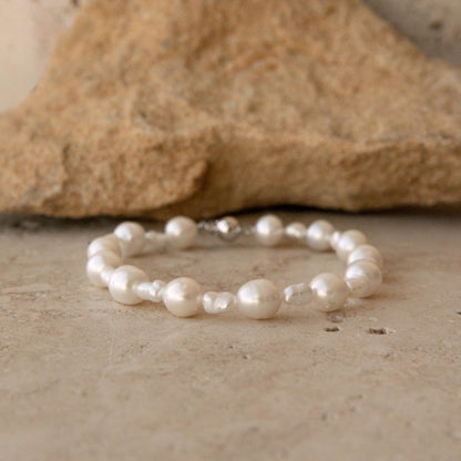 A pearl strand bracelet made of white baroque and Keshi pearls, displayed on a textured stone surface with a larger rock in the background.