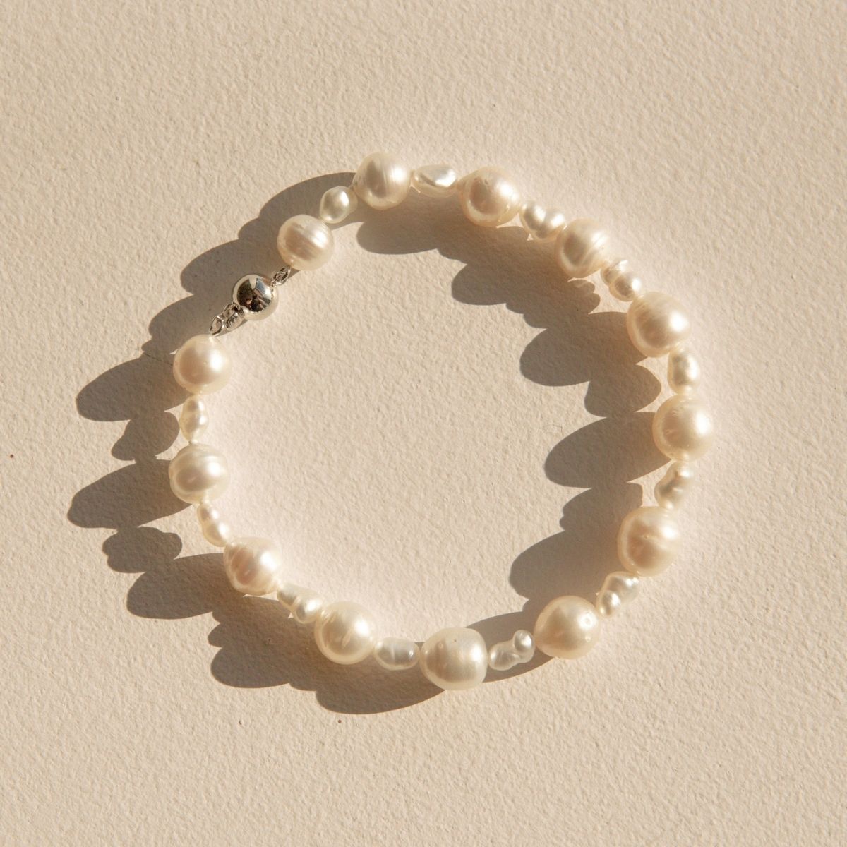 A pearl strand bracelet made of irregular Keshi and South Sea pearls, arranged in a circle on a textured beige surface with hard sunlight casting shadows.