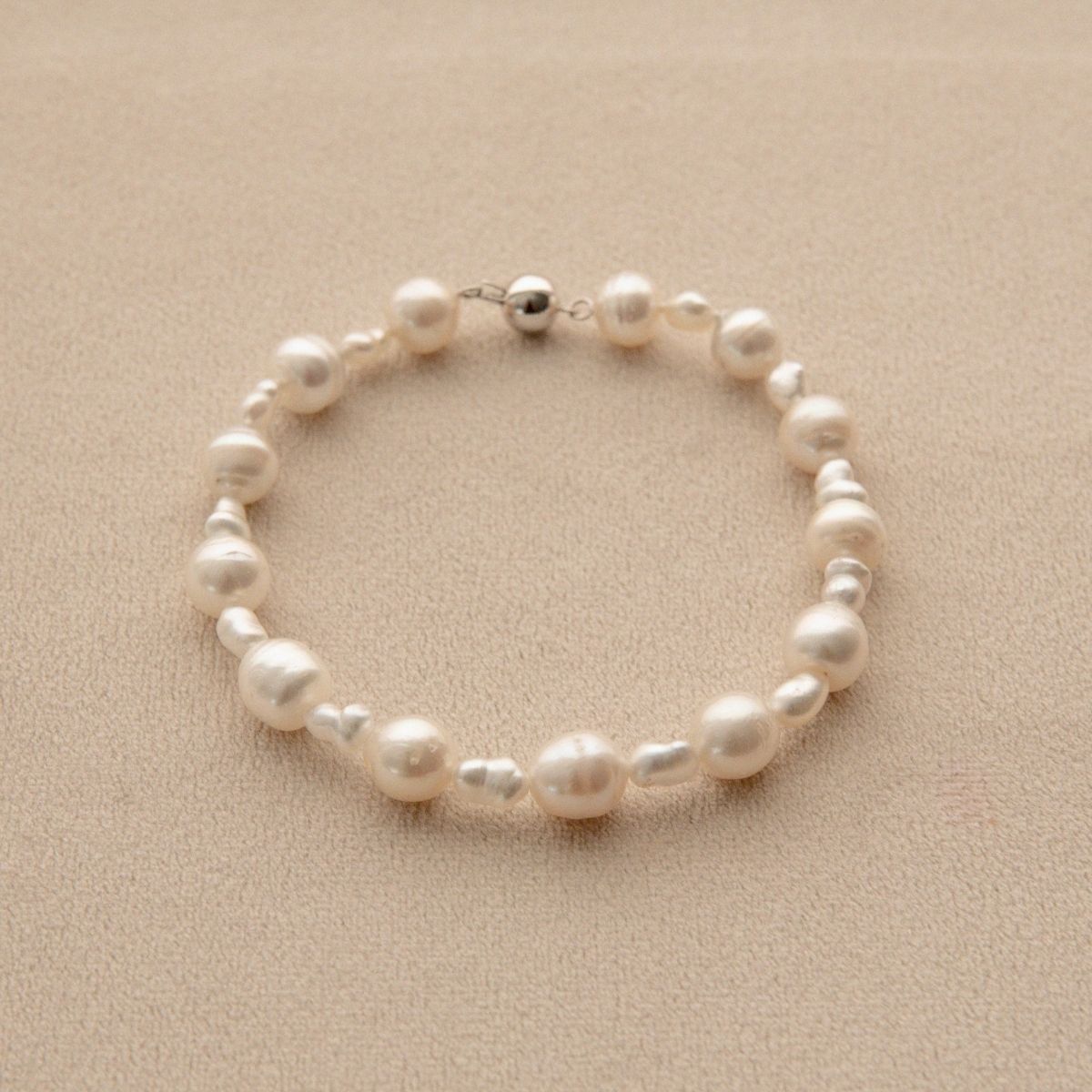 A pearl bracelet with mixed sizes of white Keshi and South Sea pearls, displayed on a textured beige surface.