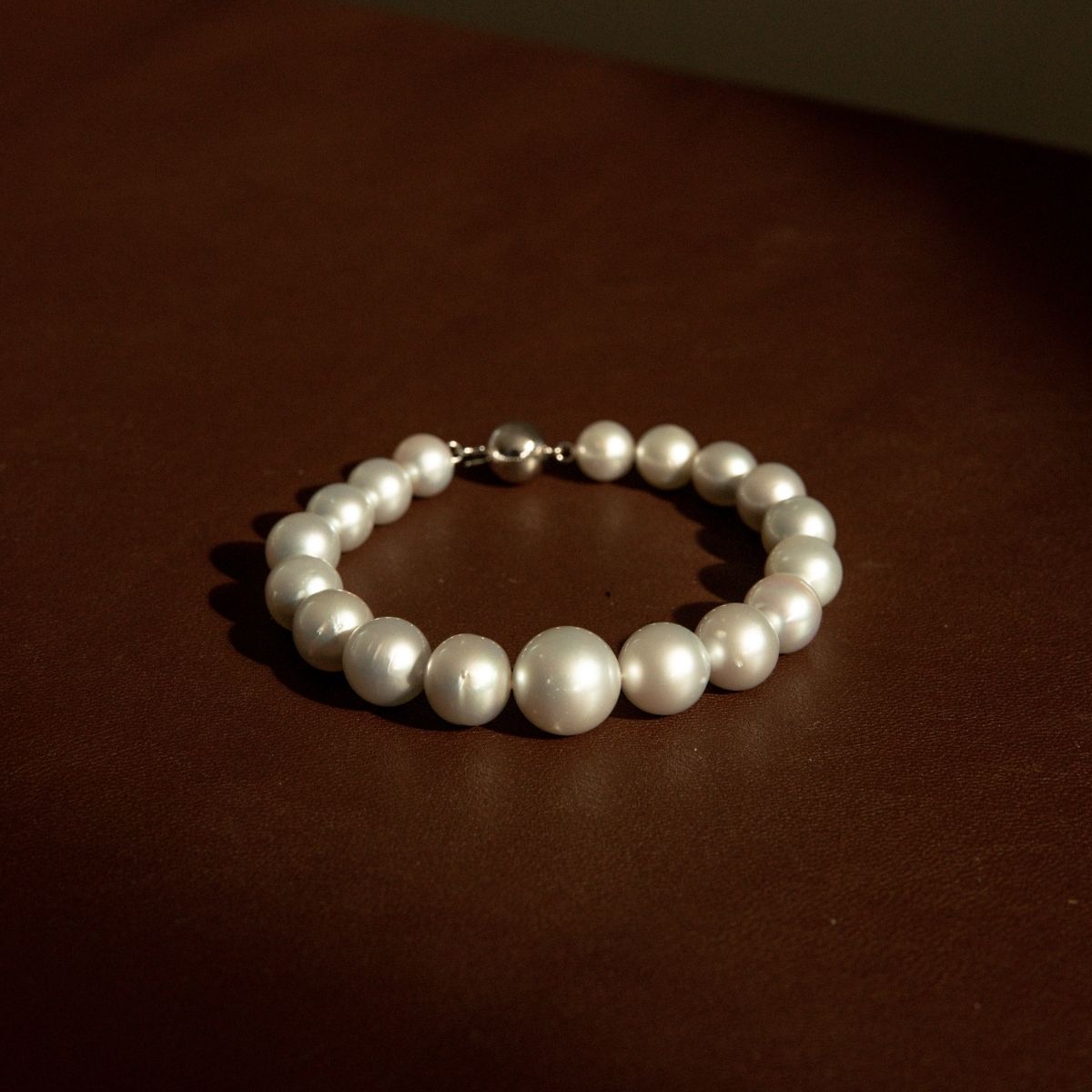 A strand bracelet made of silver South Sea pearls with a white gold metal clasp, resting on a dark brown leather surface.