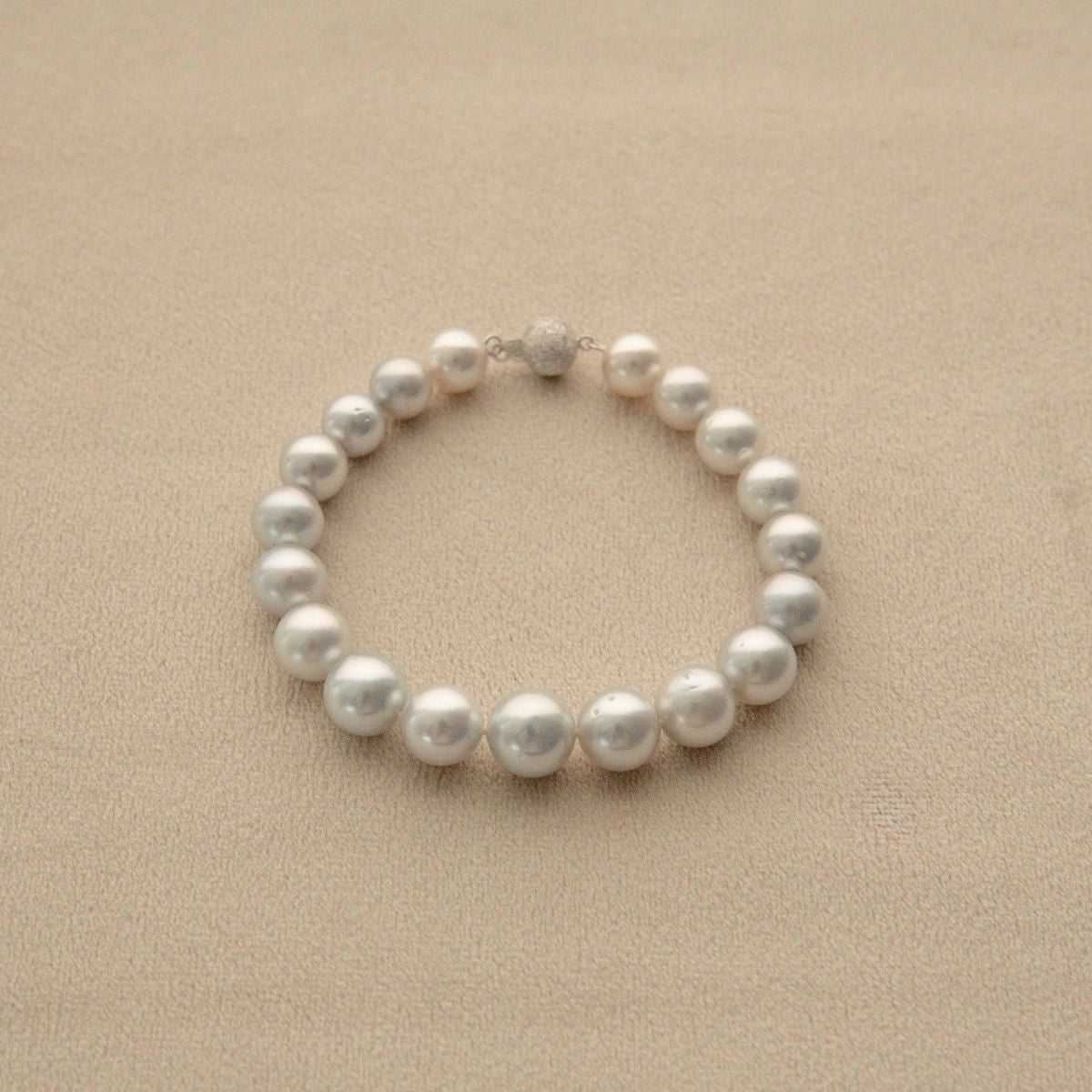 A strand bracelet made of white, round Australian South Sea pearls with a small 9ct white gold clasp, displayed on a textured beige surface.