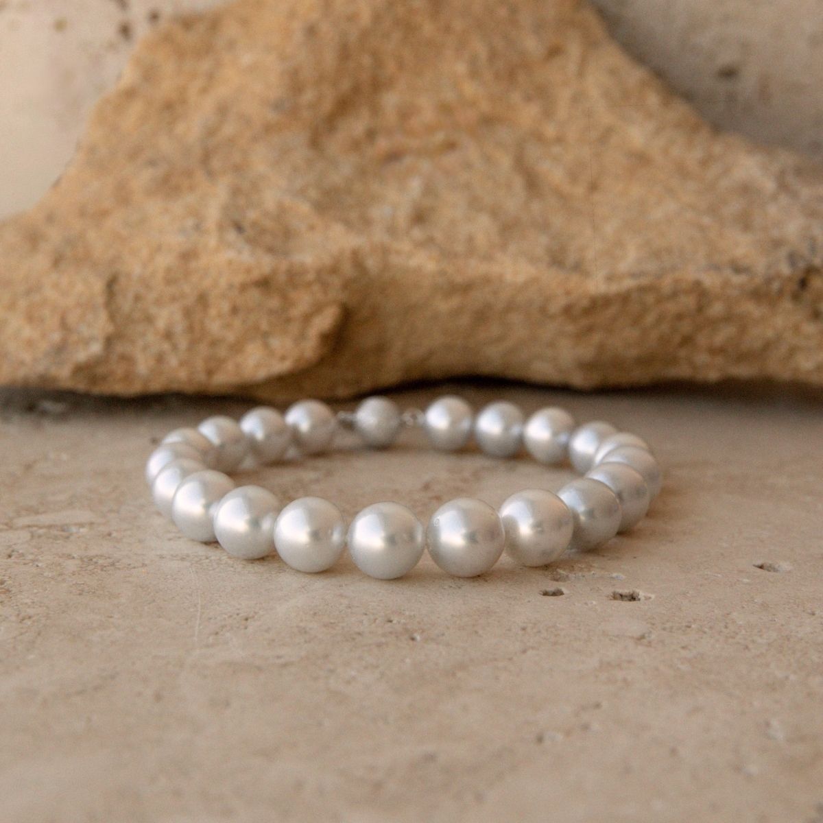 A pearl strand bracelet made of round, lustrous white South Sea pearls, displayed on a textured beige stone surface with a larger rock in the background.