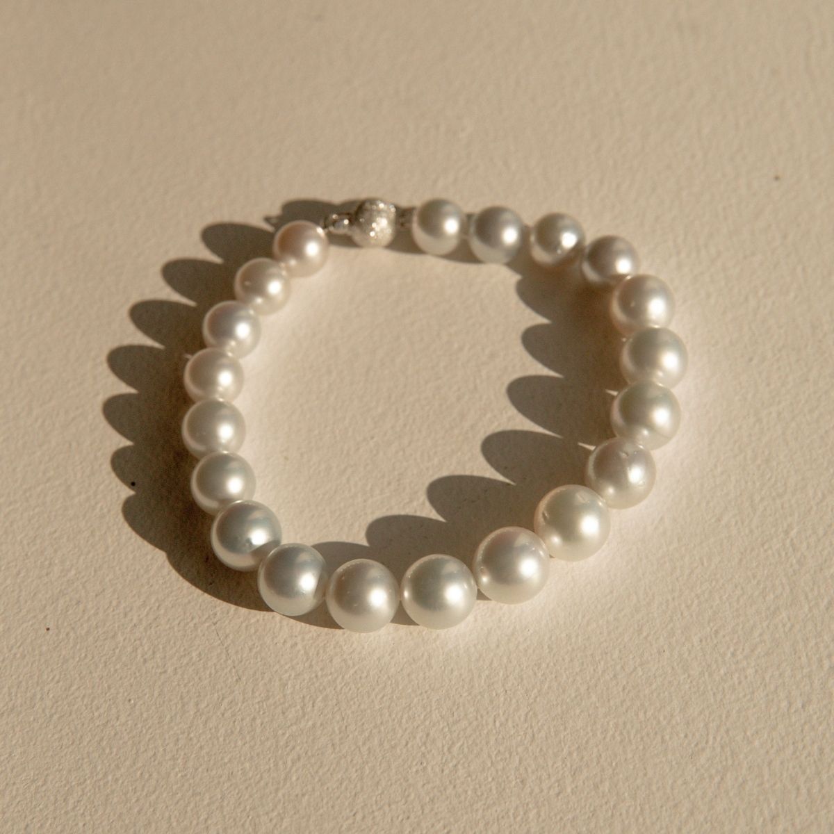 A single-strand pearl bracelet with a 9ct white gold ball clasp, arranged in a circle on a textured beige surface with soft shadows.