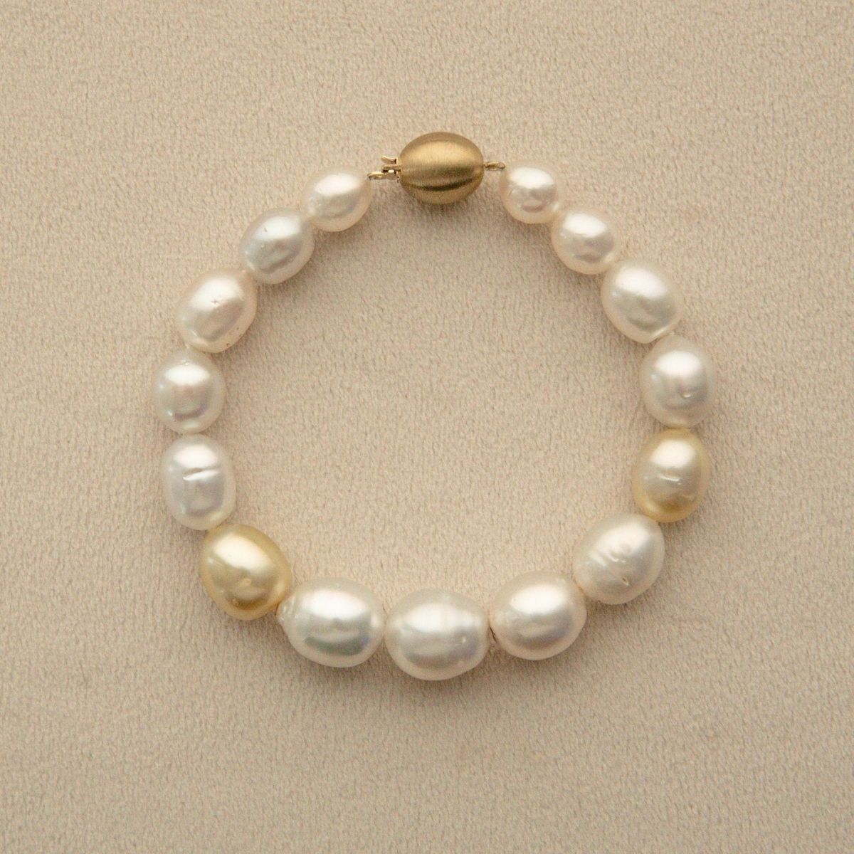 A Semi Drop pearl strand bracelet with a gold clasp, displayed on a textured beige background.