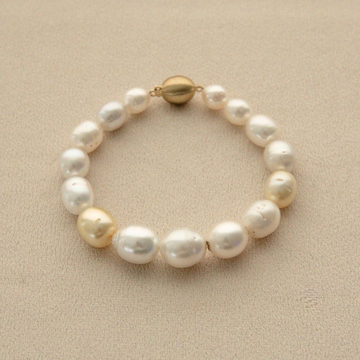 A strand bracelet made of multicolour Australian South Sea pearls with two prominent champagne-gold pearl and a gold clasp, displayed on a textured beige surface.