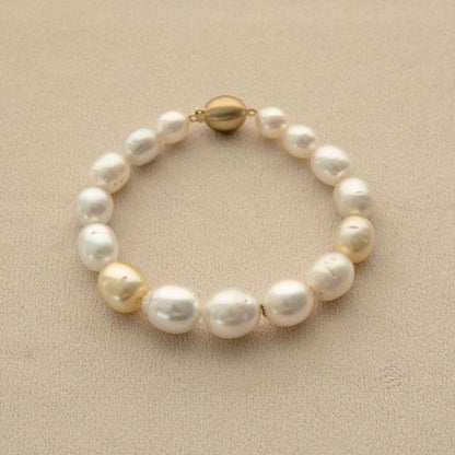A strand bracelet made of multicolour Australian South Sea pearls with two prominent champagne-gold pearl and a gold clasp, displayed on a textured beige surface.