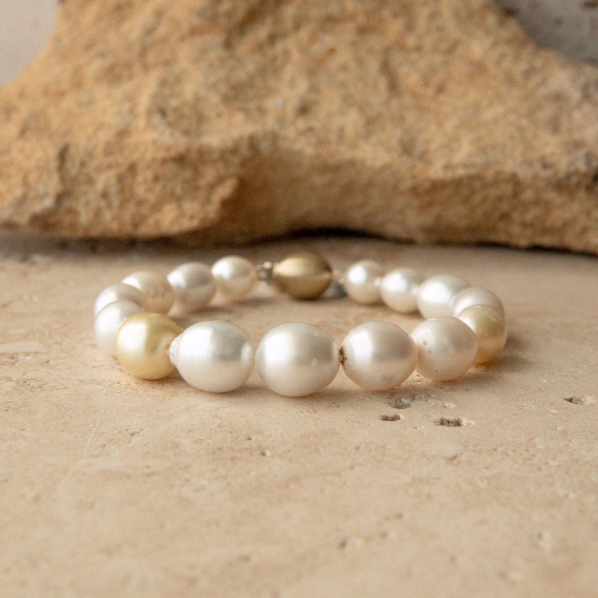 A strand bracelet made of multicolour Australian South Sea pearls with one metallic solid gold clasp, displayed on a sandy surface with a rock in the background.