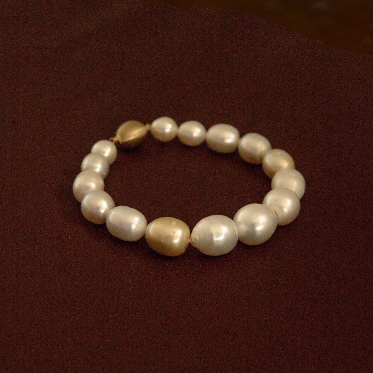 A strand bracelet made of multicolour Australian South Sea pearls with two larger champagne-gold beads, displayed on a dark reddish-brown background.
