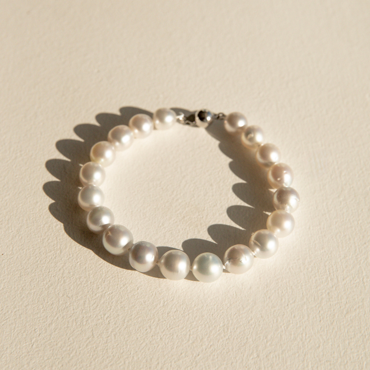 A single-strand pearl bracelet with a silver clasp, photographed on a textured beige surface with a sharp shadow.