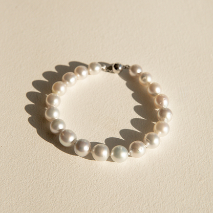 A single-strand pearl bracelet with a silver clasp, photographed on a textured beige surface with a sharp shadow.