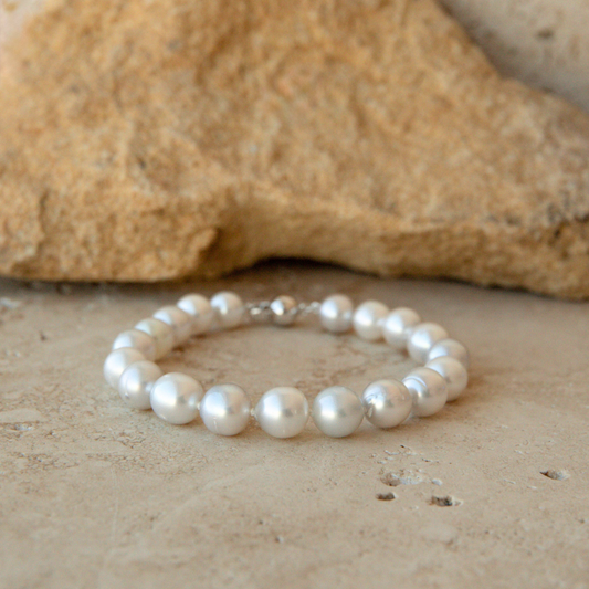 A strand bracelet made of stunning white Australian South Sea pearls with a silver clasp, resting on a textured beige stone surface with a larger tan rock in the background.