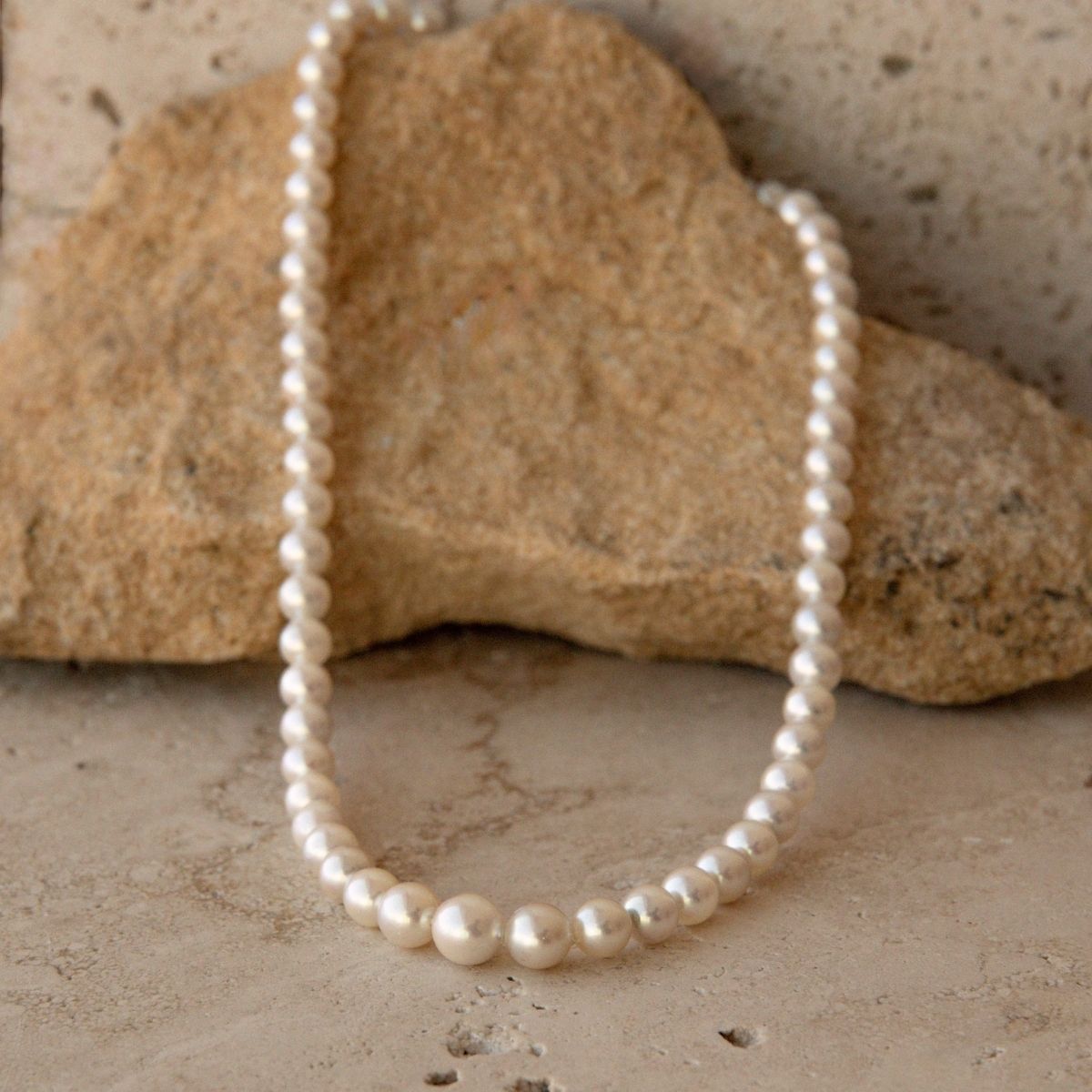 A strand of white Akoya pearls draped over a natural tan rock on a textured stone surface.