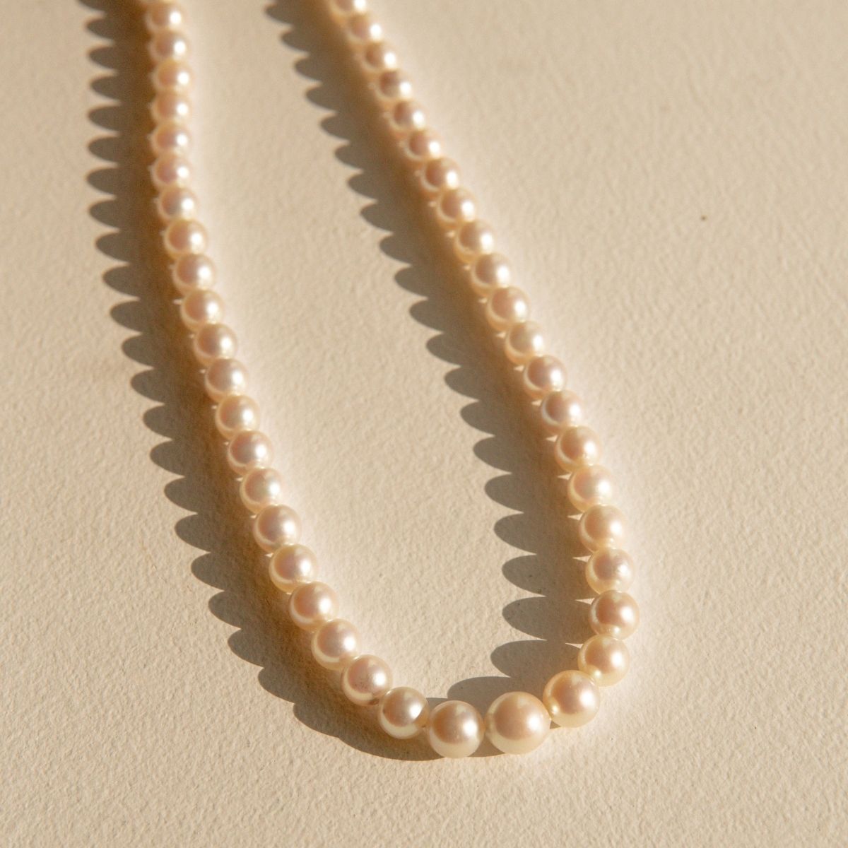 A strand of creamy off-white Australian Akoya pearls arranged in a loose loop on a textured beige surface, casting soft shadows.