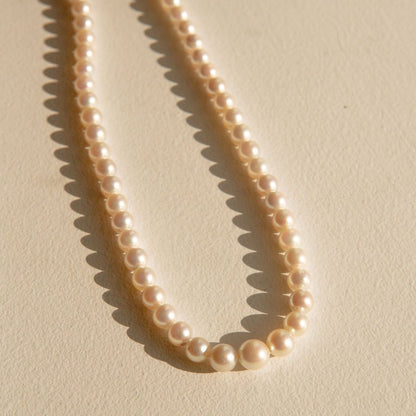 A strand of creamy off-white Australian Akoya pearls arranged in a loose loop on a textured beige surface, casting soft shadows.