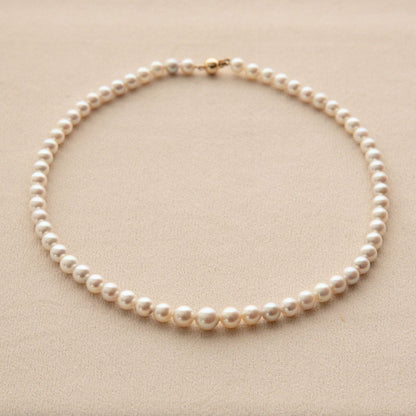 A single-strand Akoya pearl necklace with a 18ct yellow gold clasp, arranged in a circle on a textured beige background.