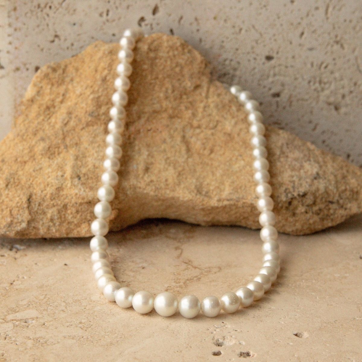 A strand of white Australian Akoya pearls arranged in a loop, resting against a natural tan rock on a textured stone surface.
