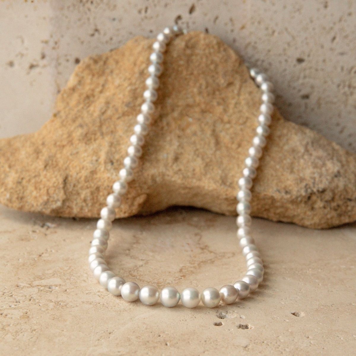 A white Australian Akoya pearl strand necklace draped over a natural tan rock on a textured beige stone surface.