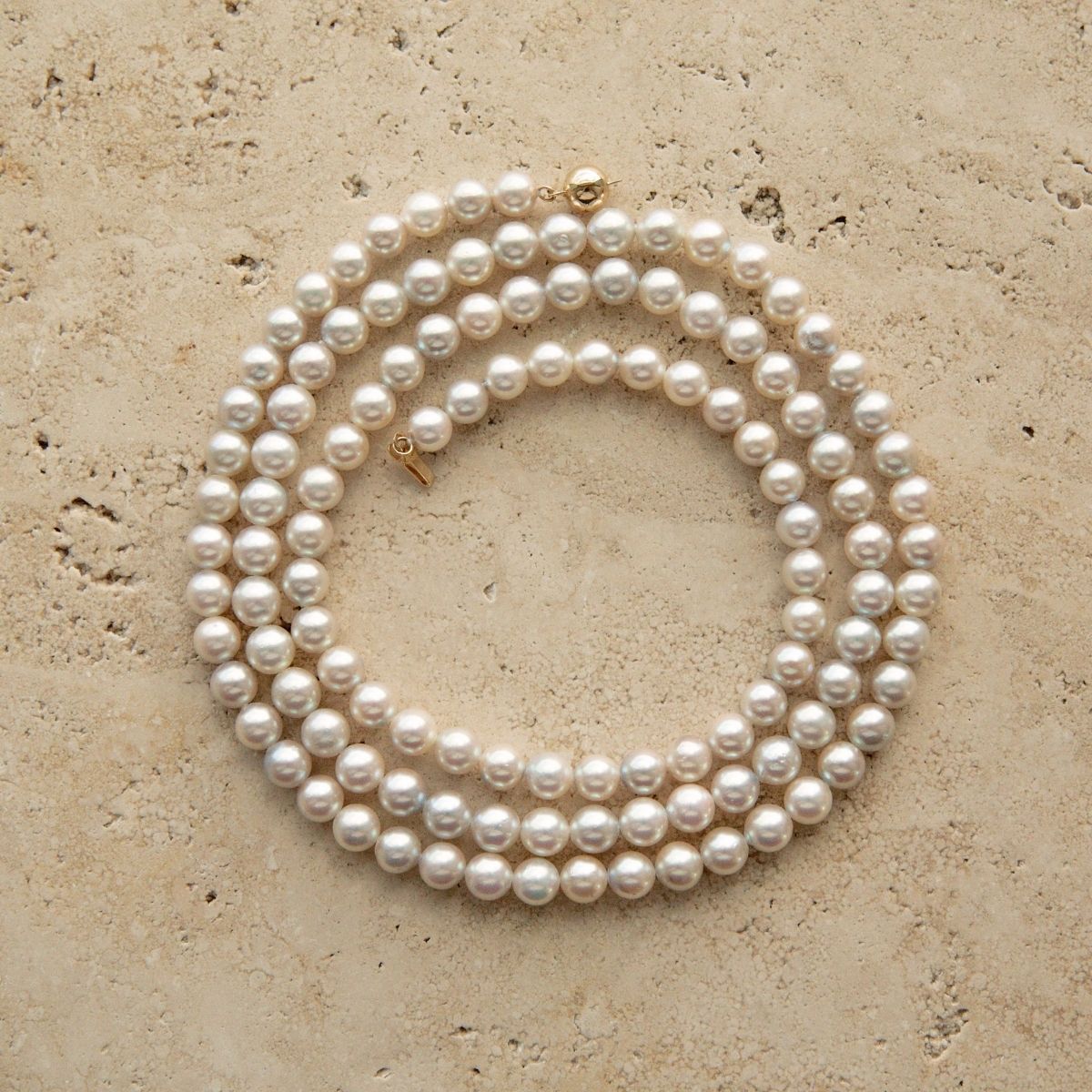 A long Australian Akoya pearl strand necklace coiled in a circle on a textured beige stone surface, featuring a small gold clasp.