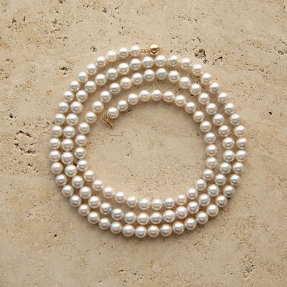 A long Australian Akoya pearl strand necklace coiled in a circle on a textured beige stone surface, featuring a small gold clasp.
