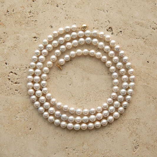 A long Australian Akoya pearl strand necklace coiled in a circle on a textured beige stone surface, featuring a small gold clasp.