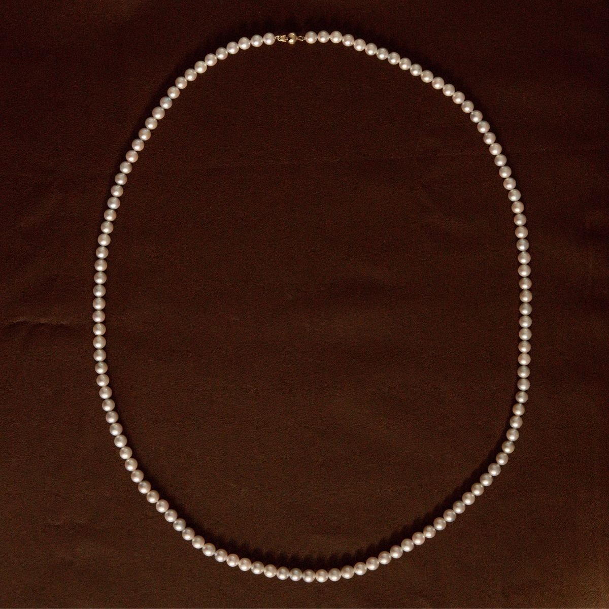 A classic Akoya pearl necklace arranged in a neat oval shape on a dark brown textured fabric background.