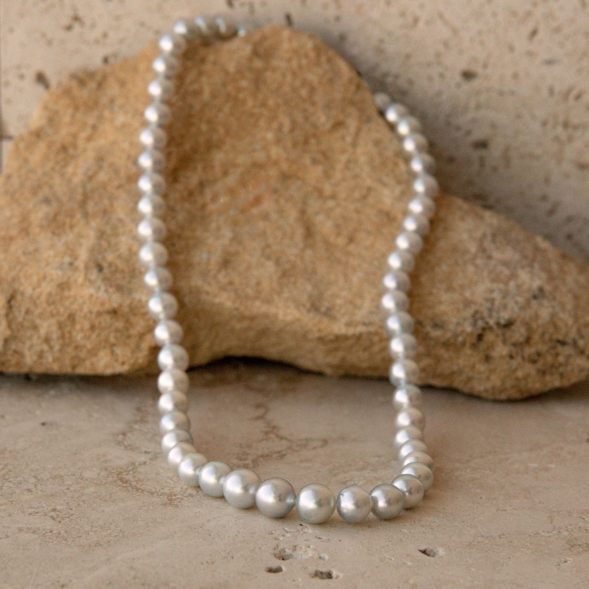 A strand of silver toned Australian AKoya pearls draped over a tan rock on a textured stone surface.