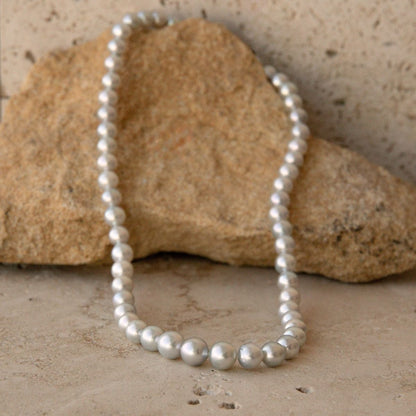 A strand of silver toned Australian AKoya pearls draped over a tan rock on a textured stone surface.