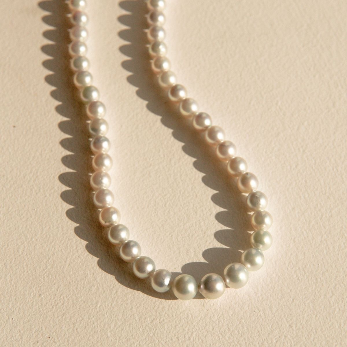 A strand of silver Australian Akoya pearls arranged in a loose loop on a textured beige surface, casting soft shadows.