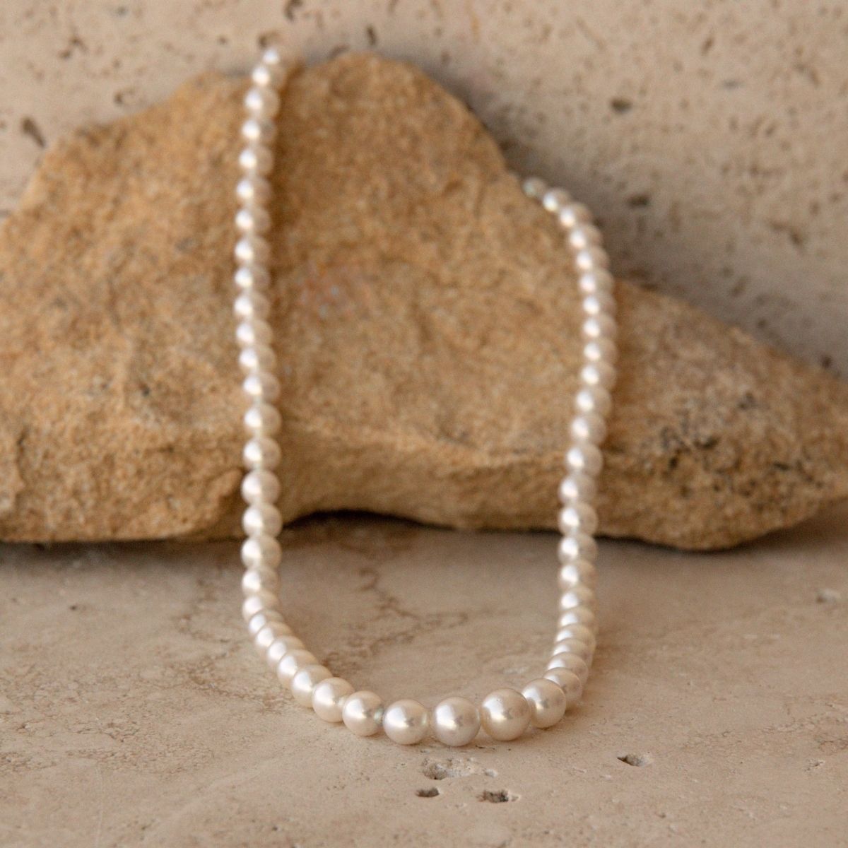 A strand of white Australian Akoya pearls draped over a natural tan rock, set against a textured beige stone background.