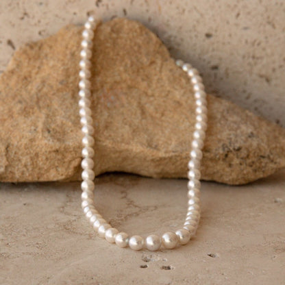 A strand of white Australian Akoya pearls draped over a natural tan rock, set against a textured beige stone background.